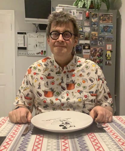 Reviewer wearing the Disney Ratatouille 'Bon Rat-Pétit' shirt, sitting at a dining table with an empty plate.