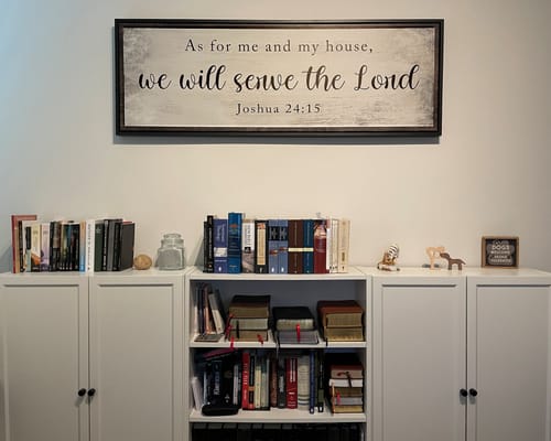 Reviewer's framed Joshua 24:15 sign mounted on an office wall above a white bookcase filled with books.