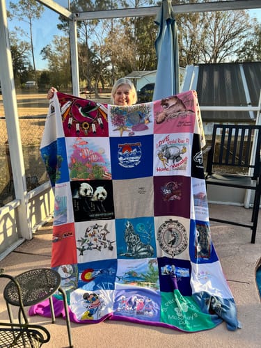 A reviewer holds up her large, colorful Premium T Shirt Quilt made from various shirts on a patio.