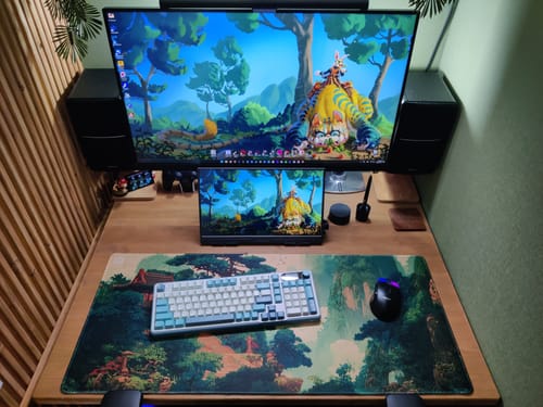 Customer's Rainforest desk pad on a wooden desk, part of a dual-monitor computer setup with a keyboard and mouse.