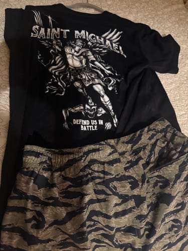 Reviewer's Saint Michael Defend Us In Battle Tee and camouflage shorts laid out on a bed.