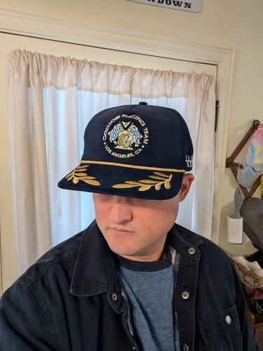 Reviewer wearing the dark corduroy Big Racing Hat with gold embroidered details on the bill.