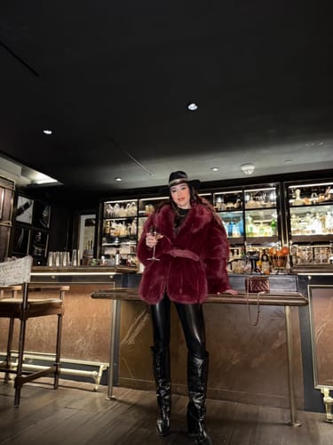 Reviewer in a bar wearing the plush, burgundy Faux Mink Pelted Coat with a matching belt, black pants, and boots.