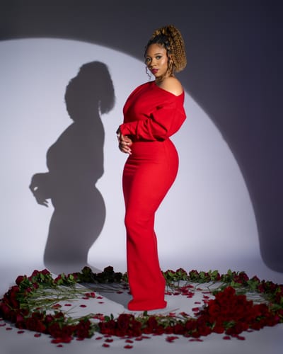 Customer in a studio photoshoot wearing the red Knit Off-Shoulder Sweater Maxi Dress, standing in a circle of roses.