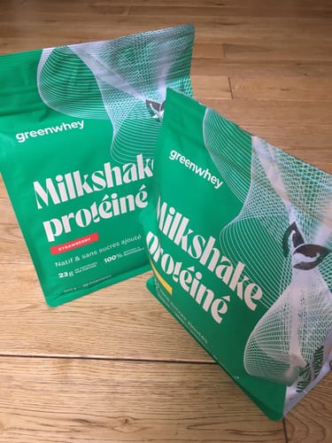 Photo d'un client montrant deux sachets de Milkshake protéiné, un à la fraise et l'autre, probable mangue, sur un sol en bois.