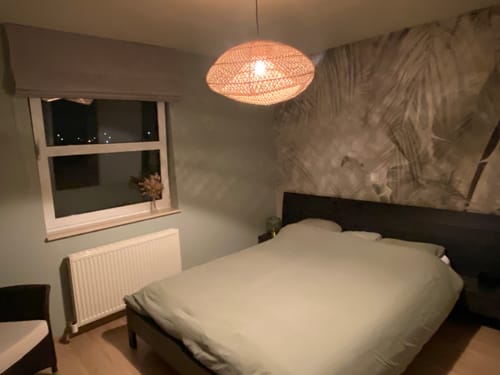 Recensent toont de rotan Maze Hanglamp die brandt boven een bed in een slaapkamer.