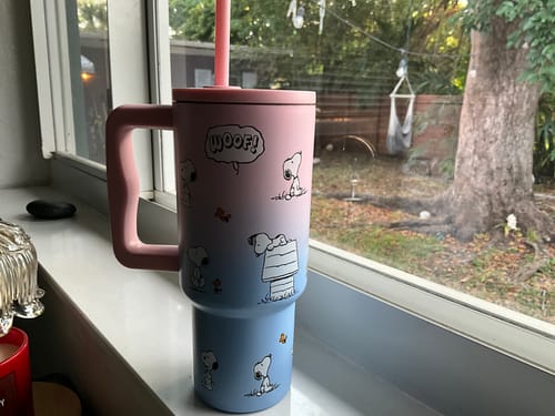 Customer's ombré Snoopy Woof Travel Tumbler with its pink handle and lid, shown on a windowsill.