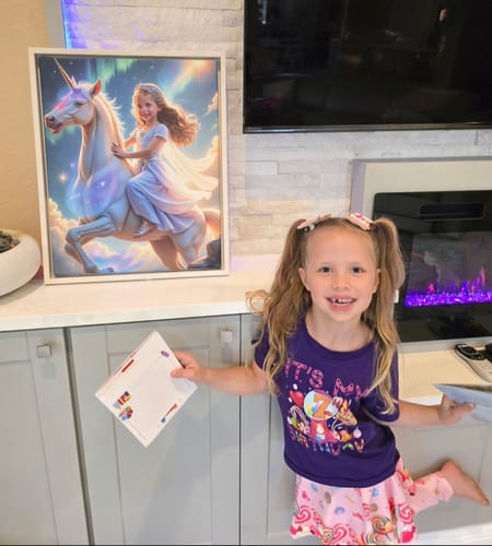 A smiling child stands next to her framed Aurora Unicorn Princess portrait displayed on a counter.