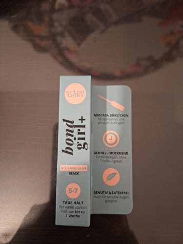 Kunde zeigt die Verpackung des BOND GIRL+ Wimpernklebers schwarz neben der Karte mit den Produktmerkmalen.