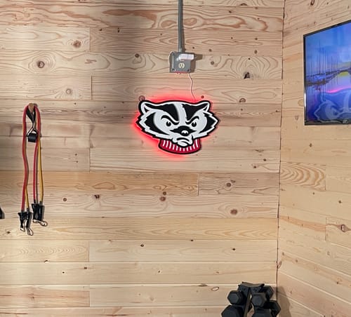 Reviewer's Wisconsin Badgers Mascot Head art with a red LED backlight, mounted on a wood-paneled wall in a gym.