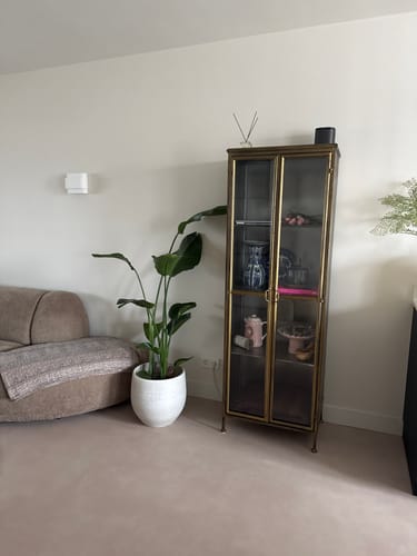 Klantfoto van de goudkleurige Gertlush vitrinekast, opgesteld in een woonkamer naast een bank en een grote plant.