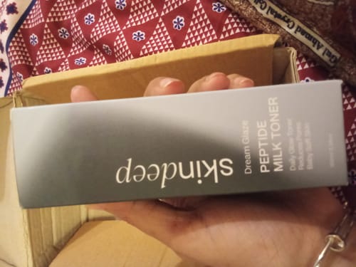 A reviewer holds the grey and white box of DREAM GLAZE Peptide Milk Toner in their hand.