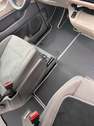 Recenzent pokazuje czarne dywaniki samochodowe z jasną lamówką, dopasowane do przedniej części vana.