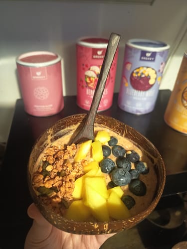 Ein Kunde hält eine Protein Choco Himbeere Bowl in einer Kokosnussschale, garniert mit Mango, Blaubeeren und Müsli.