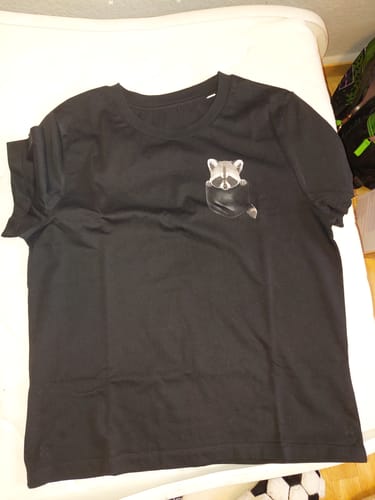 Ein Kunde zeigt das schwarze 'Waschbär Tasche' Bio-Damen-Shirt, das flach auf einer Oberfläche liegt.