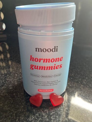 Reviewer's light blue bottle of Moodi Hormone Gummies with two red, heart-shaped gummies on a counter.