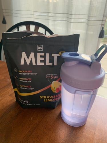 A customer's pouch of Strawberry Lemonade Melt supplement next to a purple shaker bottle on a wooden table.
