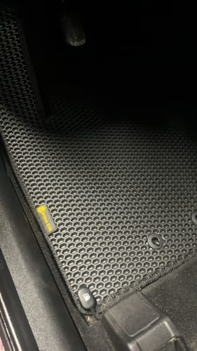 Recenzent pokazuje czarny, dopasowany dywanik samochodowy ze strukturą plastra miodu, zamontowany w aucie.