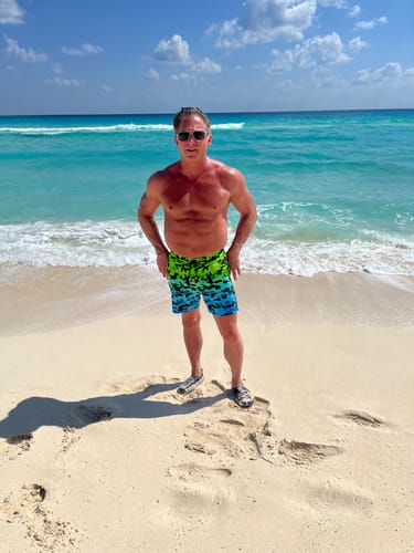Reviewer standing on a tropical beach wearing the Faded Camo - Jungle 5" swim trunks.