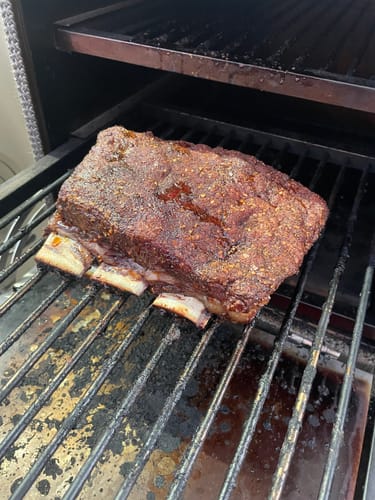 Customer's large rack of seasoned Bone-In Short Ribs cooking slowly on a smoker.