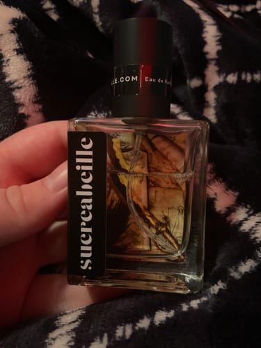 Reviewer holding the clear, square bottle of Swashbuckler perfume against a dark, fuzzy surface.