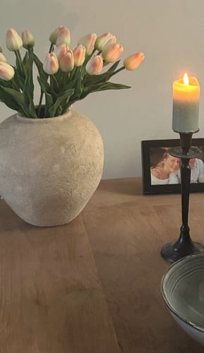 Klant toont de beige Arda vaas, gevuld met een boeket lichtroze tulpen op een houten tafel.