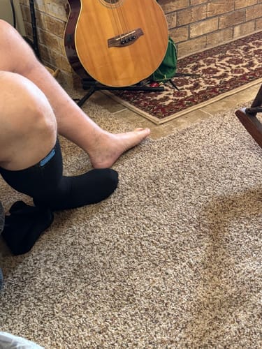 Reviewer sitting on a carpeted floor, wearing one black Performance Eucalyptus Compression Sock.