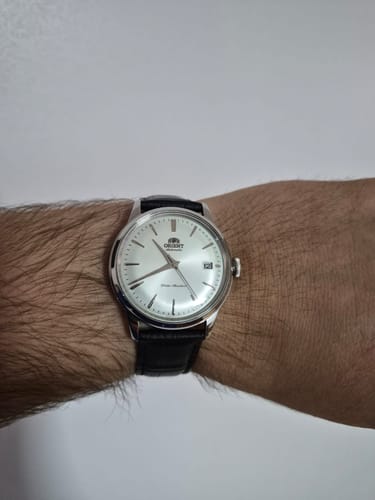 El cliente muestra su elegante reloj Orient Bambino en la muñeca, con esfera clara y correa de cuero negra.