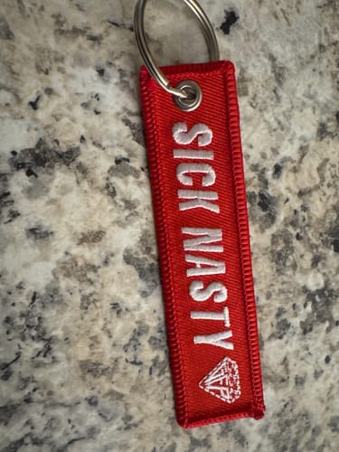 Customer's red SICK NASTY KEYTAG with white embroidered lettering lying flat on a mottled countertop.