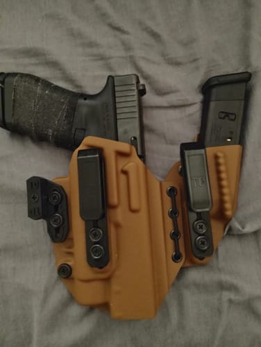 A reviewer's tan AXIS ELITE holster holding a black handgun with a taped grip and a spare magazine, lying on a gray surface.
