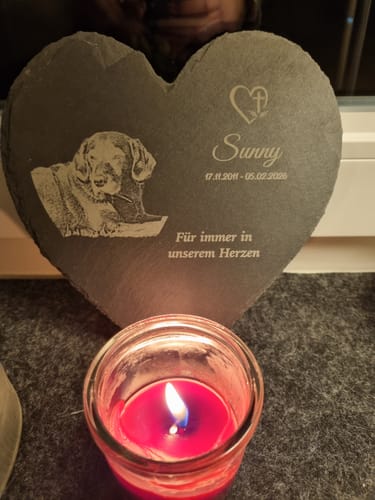 Ein Kunde zeigt das personalisierte Schieferherz mit Gravur eines Hundes und dem Namen 'Sunny' vor einer brennenden Kerze.