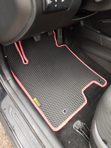 Recenzent pokazuje idealnie dopasowany czarny dywanik samochodowy z różową lamówką po stronie kierowcy.