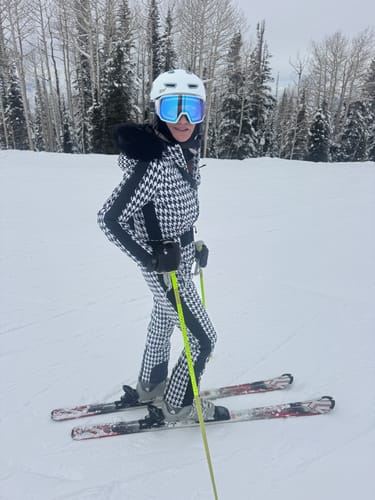Reviewer wearing the slim-fit houndstooth one-piece ski suit with its faux fur hood on a snowy slope.