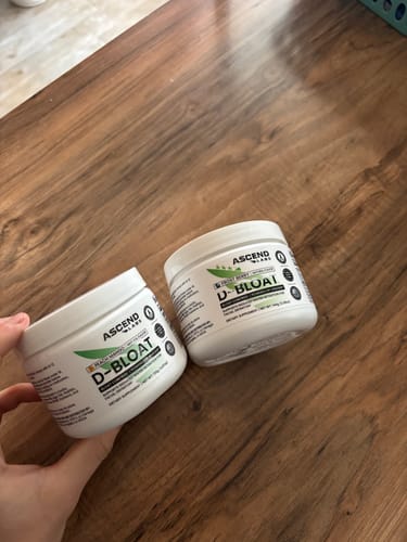 Customer holding a white tub of D-BLOAT, with a second tub resting on a wooden table.