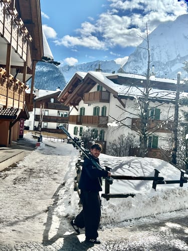 Reviewer walking through a snowy mountain village wearing the black snow jacket and carrying skis.