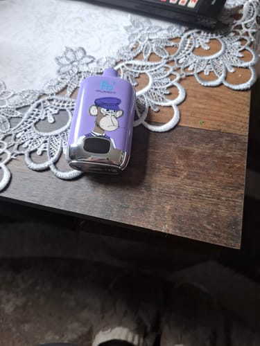 Ein Kunde zeigt die lila RANDM DIGITAL BOX 12000 Vape mit Affen-Design, die auf einem Holztisch liegt.