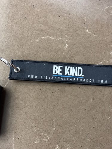 Reviewer's black Be Kind flight tag with white 'BE KIND.' text, shown lying on a surface.