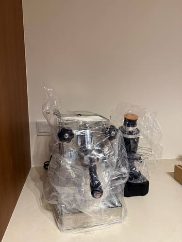 Customer's new chrome espresso machine and grinder in protective wrapping on a kitchen counter.