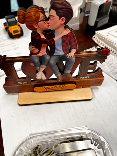 Customer's personalized wooden 'LOVE' plaque showing a kissing couple, with the names 'Beth' and 'James', on an office desk.
