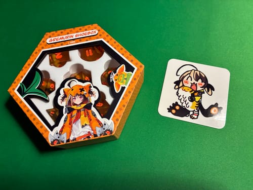 Customer's Gigi Murin Resin Dice Set in its orange hexagonal box, next to a character sticker on a green surface.