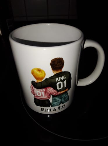 Ein Kunde zeigt eine personalisierte, weiße Tasse mit einem Paar von hinten und dem Aufdruck 'King 01' und 'Queen 01'.