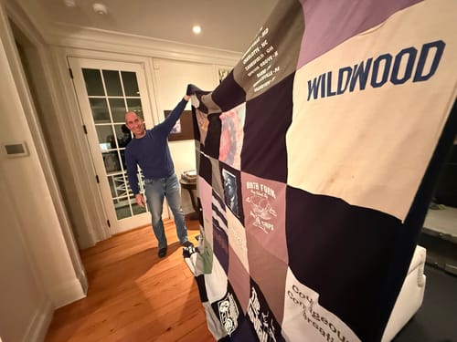 Reviewer smiling while holding up their large Premium T Shirt Quilt inside their home.