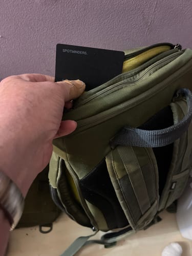 Reviewer placing the black SPOTMINDERS Tracking Card into the pocket of a green backpack.