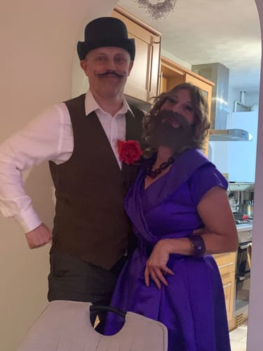 Two customers in fun circus costumes, one with a fake beard and the other with a mustache, playing the murder mystery game.