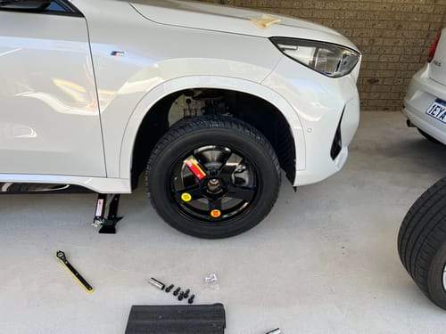 Customer's black Space Saver Spare Wheel Kit fitted onto a white BMW iX1, which is raised on a jack in a garage.