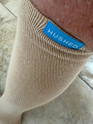 Close-up of a customer wearing the tan compression sock, showing the ribbed top band with a blue 'HUSHED' logo.