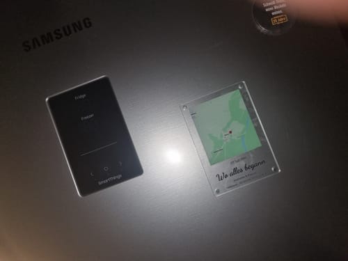 Rezensent zeigt den klaren 'Unser Ereignis' Acrylmagneten mit einer Karte an einem Kühlschrank.