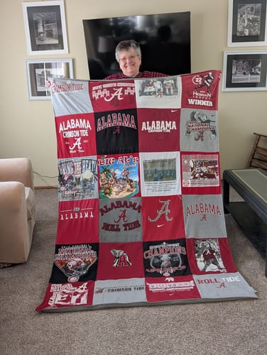 Customer holding up a large Premium T Shirt Quilt made from red and grey Alabama-themed t-shirts in her living room.