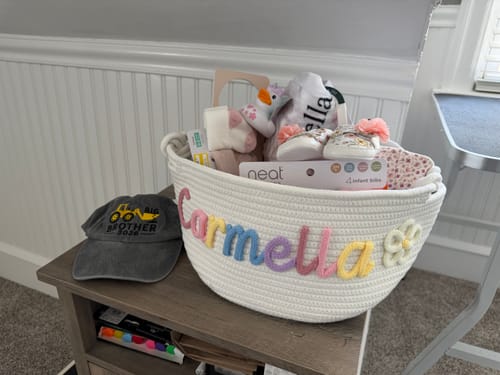 Customer's personalized knitted basket with "Carmella" in multicolor letters and a flower, filled for a baby shower.