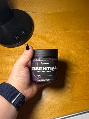 Reviewer holding a black jar of Essentials Multivitamin against a wooden surface.